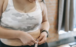 Young obese female using tape for evaluating size of belly while preparing for training in modern fitness center
